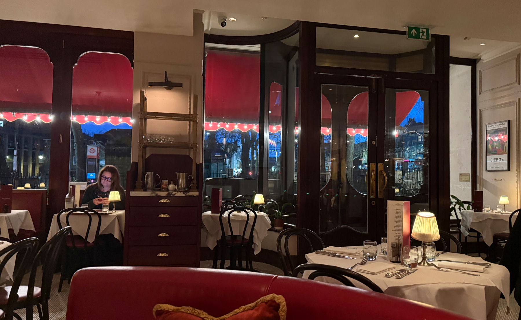 A cozy restaurant interior features red accents, round tables set for dining, and a woman seated alone near the window.